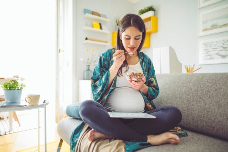 A pregnant woman sits cross-legged on a couch, eating cereal from a bowl and looking at a paper in her lap—likely related to her pregnancy diet. She is dressed in casual clothes with a blue patterned robe. The room features a light interior with shelves, plant decor, and framed pictures.