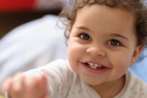 A toddler with curly hair smiles brightly while looking slightly upward. The child, perhaps imagining a name like Julia or Jasmine, is wearing a white long-sleeve shirt with a subtle pattern. The background is blurred, featuring soft colors and indistinct shapes.