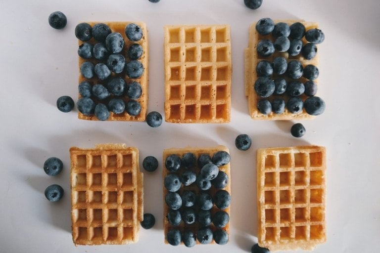 Six rectangular waffles arranged in two rows with three in each row. Five of the waffles are topped with blueberries, while one in the center is plain. Scattered blueberries are also visible on the white surface surrounding the waffles, making for delightful breakfast ideas.
