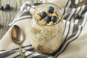 A jar filled with overnight oats, topped with blueberries, rests on a white and black striped cloth next to a silver spoon. The table surface appears to be wood. Perfectly crafted for those seeking best foods for breastfeeding, this nutritious delight is both wholesome and delicious.