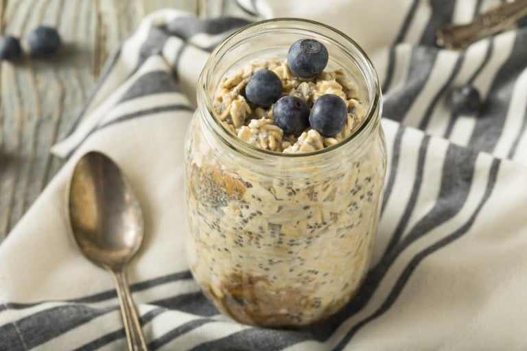 A jar filled with overnight oats, topped with blueberries, rests on a white and black striped cloth next to a silver spoon. The table surface appears to be wood. Perfectly crafted for those seeking best foods for breastfeeding, this nutritious delight is both wholesome and delicious.