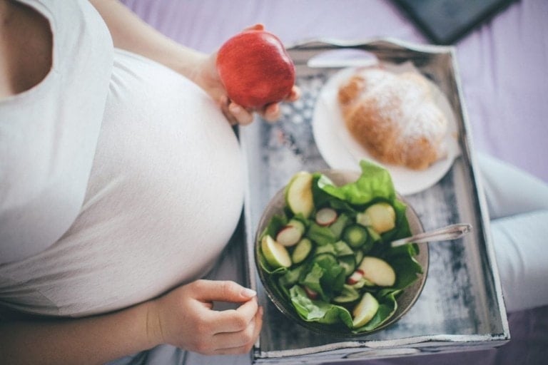 A pregnant person is holding an apple while sitting with a tray of food resting on their lap. The tray includes a bowl of salad with greens and sliced vegetables, alongside a plate with a croissant—a perfect array of foods to eat while pregnant.