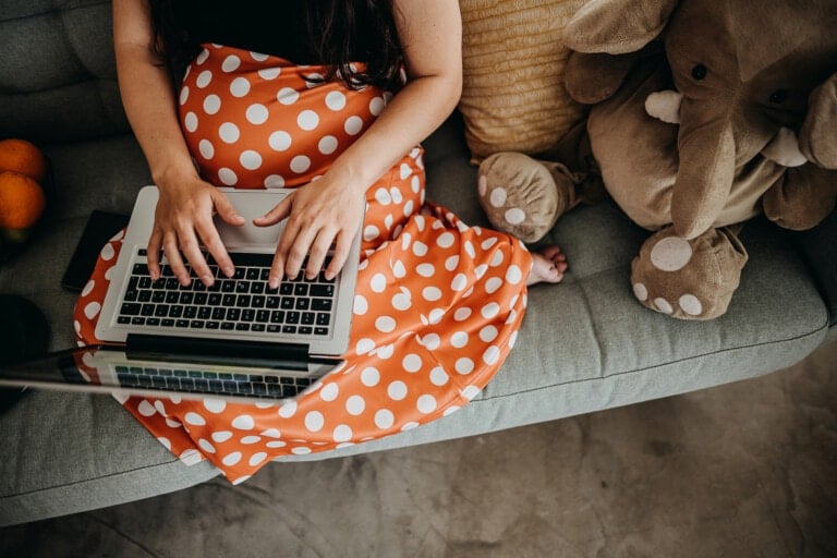 A person sits on a couch working on a laptop resting on their lap. They are wearing an orange and white polka dot dress. Beside them are an elephant stuffed animal and a pillow, possibly items from an Amazon baby registry review. An orange object is partially visible on the left side of the image.