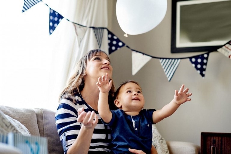 A woman and a child, both smiling, are sitting on a couch. The child is reaching up towards a white balloon in the air. Behind them, decorative blue and white triangular flags hang, creating the perfect atmosphere for a kid's birthday party. The room appears bright with natural light.
