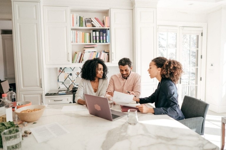 Three people are sitting at a kitchen island. One person is holding papers, while another works on a laptop. Books and colorful binders are on shelves in the background. The setting is bright and well-lit with natural light from windows, ideal for discussing whether to hire a family financial planner.