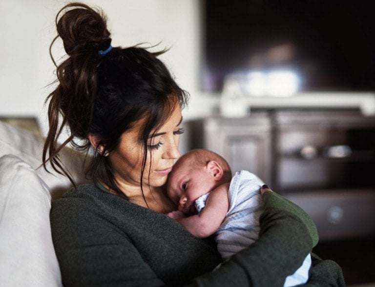 A woman with dark hair in a messy bun is sitting on a couch, holding a sleeping baby wrapped in a white blanket. They are both in a cozy indoor setting with wooden furniture and a dark television screen in the background, her peaceful expression contrasting with the occasional scary thoughts postpartum mothers often face.
