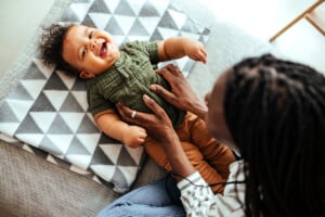 A baby wearing a green shirt and brown pants is lying on a geometric-patterned blanket, smiling and looking up. An adult, whose face is not visible, gently holds the baby’s sides while sitting on a couch, likely celebrating 5-month milestones such as increased curiosity and interaction.