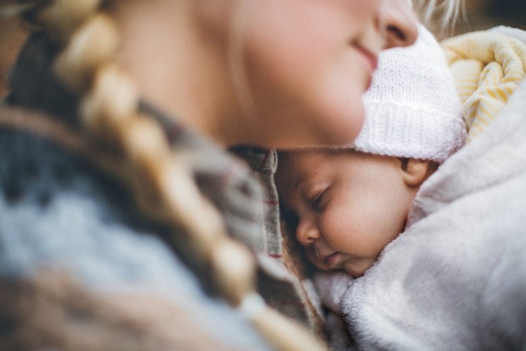 A close-up image shows a woman with a blonde braid holding a sleeping baby, dressed perfectly for winter. The baby is wrapped in a white blanket and wearing a white knitted hat. The woman's face is partially visible, looking down lovingly at the cozy little one.