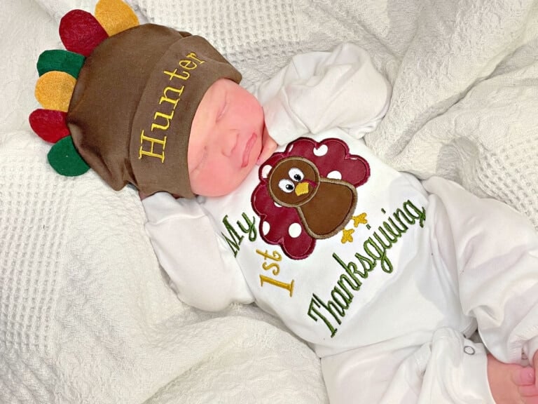 A newborn baby boy lies on a white textured blanket, wearing a charming first Thanksgiving outfit adorned with a turkey and the text "My 1st Thanksgiving." The baby also sports a brown hat with colorful feathers and the name "Hunter" embroidered on it.