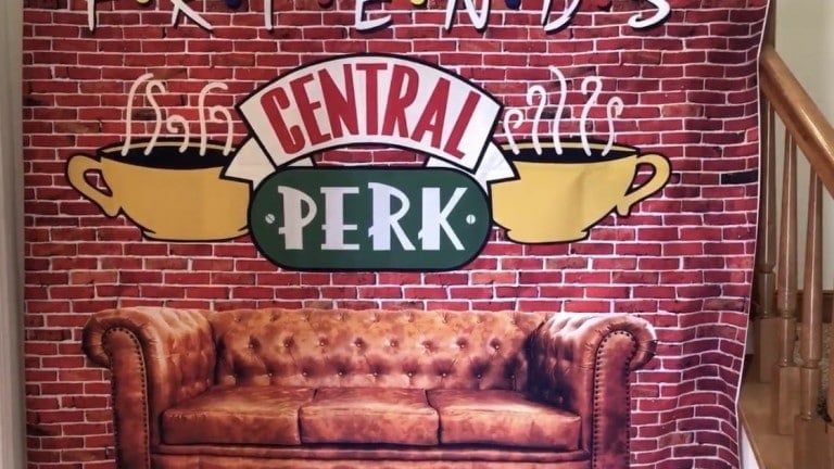 A wall hanging features a brick-patterned background with the "Central Perk" logo from the TV show "Friends." The logo is flanked by two yellow coffee cups. In front of the banner is a brown, tufted leather couch—perfect for setting the scene at a Friends birthday party.