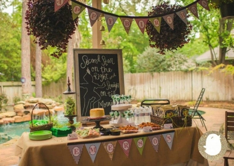 A backyard birthday bash setup featuring a table with various snacks, drinks, and treats. A chalkboard sign with Peter Pan-themed writing is visible, reading "Second star on the right and straight on till morning." Triangular banners hang above the table, creating a whimsical Neverland birthday vibe.