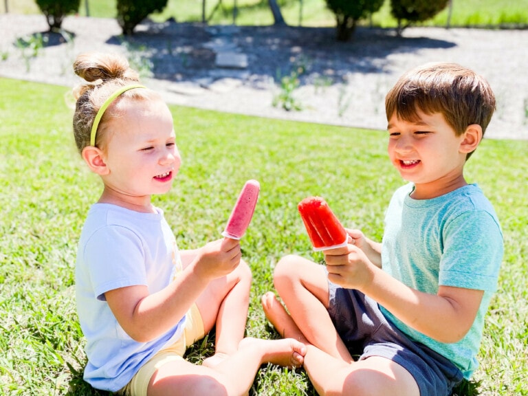 Two children are sitting on grass on a sunny day, each holding a popsicle. The child on the left is wearing a white shirt and a headband with a bun, holding a pink popsicle. The child on the right in light blue shirt and shorts holds a red popsicle. They're enjoying their treats from homemade popsicle recipes.