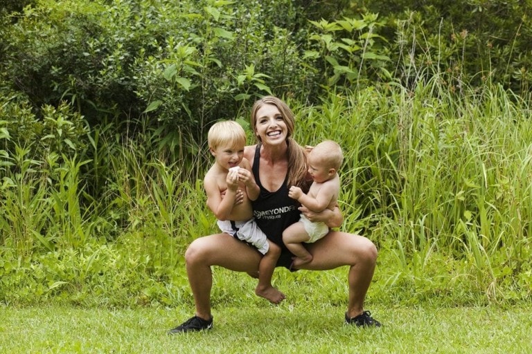 A new mom is squatting outdoors in a grassy area, holding a young child in each arm. Both children are shirtless, and the woman is smiling, wearing a black tank top and shorts. The background consists of tall grasses and greenery as she manages to find time to exercise.