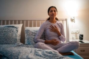 A person in pajamas sits cross-legged on a bed, eyes closed, with one hand on their chest and one on their abdomen. A lit lamp, a glass of water, and an alarm clock are on the nightstand beside the bed.