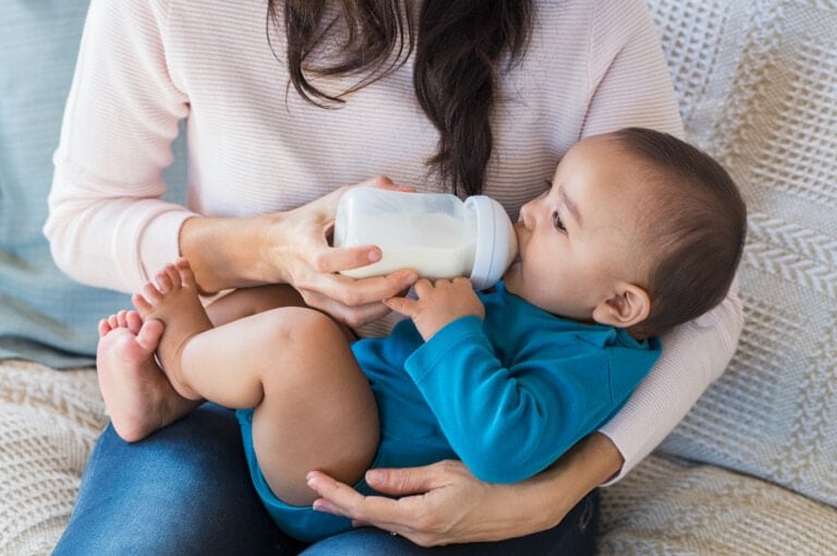 An adult practices paced bottle-feeding while holding a baby on their lap. The baby, dressed in a blue outfit, has a relaxed expression. The adult, wearing a light pink sweater, sits comfortably on a beige textured cushion.