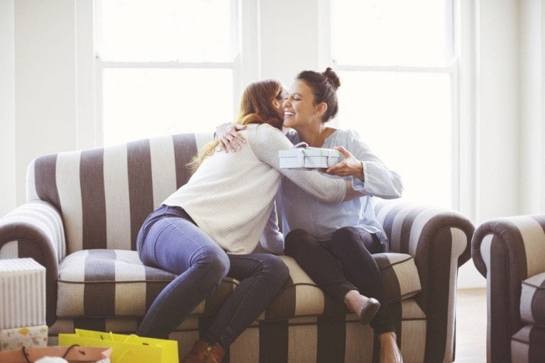 Two women sit on a striped sofa in a brightly lit room. One woman, holding a gift box, is leaning in to hug the other. Several shopping bags and other gifts—possibly the best gifts to give a pregnant mom—are visible on the floor and couch around them.