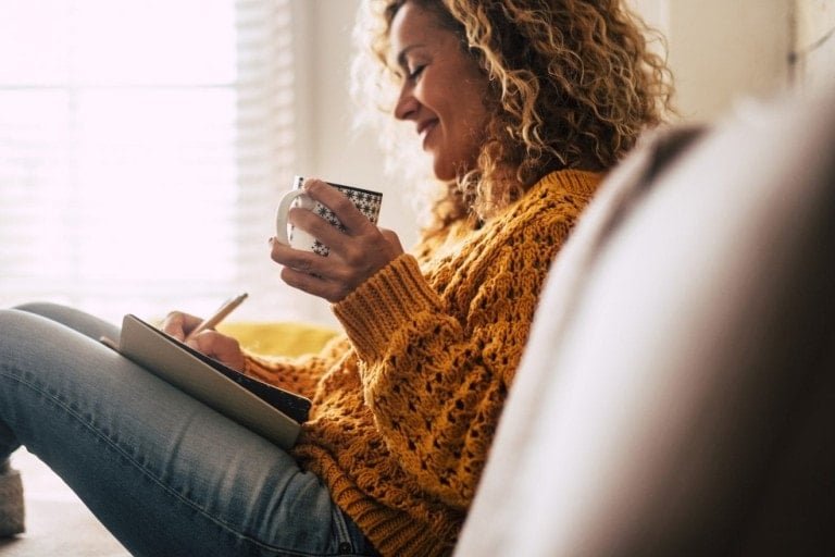 A person with curly hair sits against a wall, holding a mug in one hand and writing in a notebook with the other. They are wearing a yellow knit sweater and jeans, embodying cozy self-care essentials. Soft light enters the room through a window in the background.