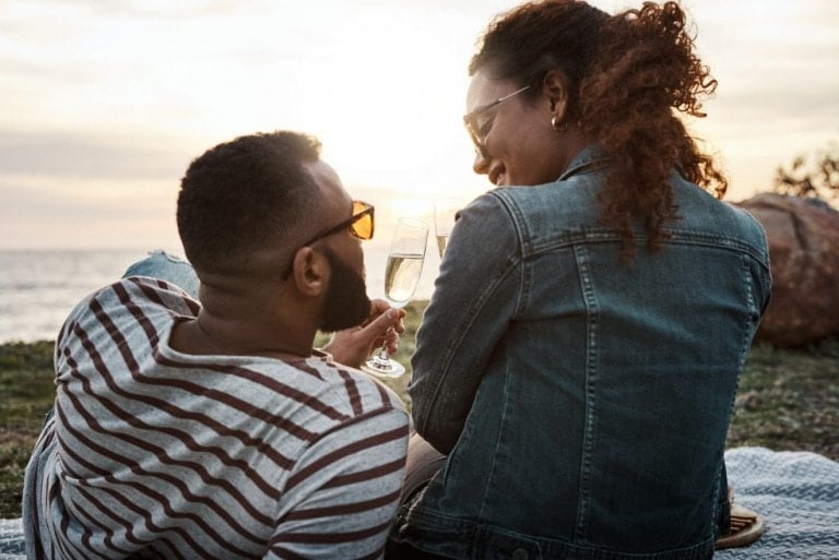 A man and a woman are sitting on a blanket at a beach during sunset, raising their glasses in a toast. The man is wearing a striped shirt and sunglasses, while the woman sports a denim jacket and sunglasses. As they stay connected with your spouse, the ocean and sun create the perfect backdrop.