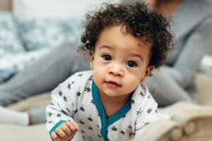 A baby with curly hair, potentially one of those adorable boy names that start with M, is dressed in a white onesie with black star patterns and teal trim, looking directly at the camera. The cozy indoor setting features a person in gray clothing visible in the blurred background.
