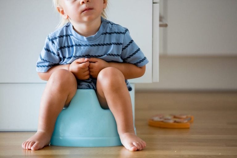 A young child with blonde hair, wearing a blue striped shirt, sits on a blue potty as they prepare for potty training. The child's face is not visible. They are barefoot on a light-colored wooden floor, with disordered toys in the background.