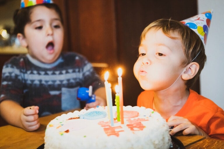 Two young children wearing party hats sit at a table with a birthday cake in front of them. One child is blowing out the candles while the other looks on with an open mouth. The cake, decorated with white frosting and three lit candles, evokes an old-school birthday charm.