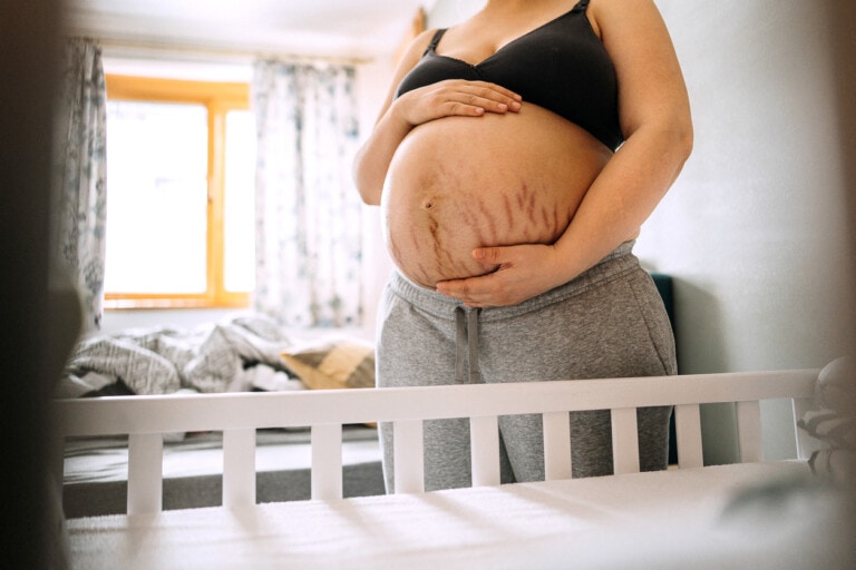A person wearing a black sports bra and gray sweatpants stands next to a white crib, holding their pregnant belly with both hands. Pregnancy stretch marks are visible on their stomach. A bed and window with curtains are in the background.