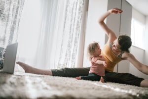 A woman in a yellow tank top and black pants is sitting on the carpet in a living room, doing stretches for mom with one leg extended. A small child in a striped shirt is stretching beside her. A laptop is placed on the floor nearby.