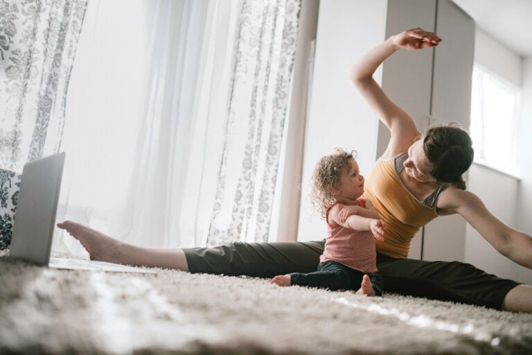 A woman in a yellow tank top and black pants is sitting on the carpet in a living room, doing stretches for mom with one leg extended. A small child in a striped shirt is stretching beside her. A laptop is placed on the floor nearby.