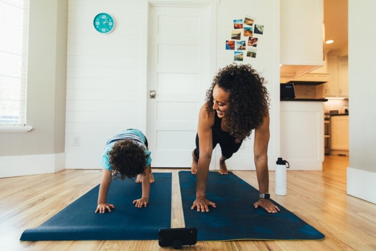 A woman and a child are engaging in a plank exercise on yoga mats in a bright room, fostering healthy habits. The woman smiles at the child as they hold the position. Nearby, there's a water bottle by the woman, a clock on the wall, photos on the door, and a smartphone placed in front of them.