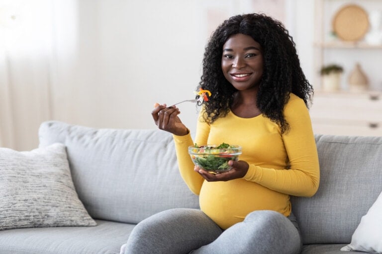 A pregnant woman wearing a yellow shirt sits on a gray couch, enjoying a bowl of salad perfect for her second trimester. She smiles, fork in hand, in the bright, cozy living area.