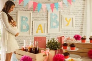 A pregnant woman in a white dress stands by a decorated table at a baby shower. The table is adorned with cupcakes, drinks, and flowers, surrounded by beautifully wrapped gifts. On the wall behind her, buntings and framed letters spell "BABY" in yellow and blue.