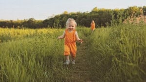 A young child wearing orange overalls runs down a grassy pathway in a green field. Another person in the distance, also clad in orange, strolls along the same path. The serene scene is set in a countryside area under a clear sky, evoking timeless country girl names.