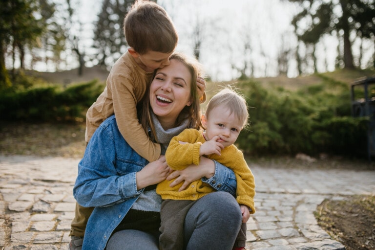 A woman sits outdoors on a cobblestone path, smiling as she embraces two young children. One child is kissing her on the forehead, and the other child is seated on her lap, looking at the camera. It's a moment that reminds her why putting her career on pause was worth it. Trees and bushes are visible in the background.