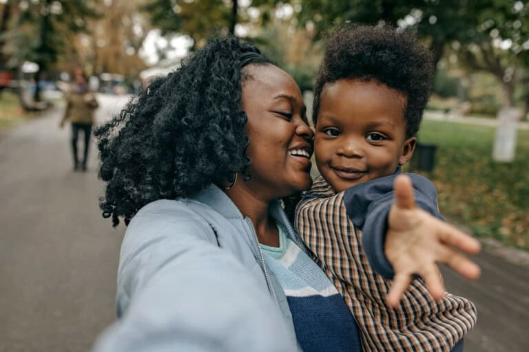 A woman holds her toddler son while smiling and taking a selfie in a park. The boy reaches towards the camera. Trees and other people walking are visible in the blurred background.