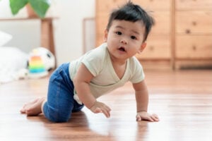 A curious 8-month-old baby is crawling on a wooden floor, wearing a light-colored shirt and blue pants. A colorful toy and wooden furniture are in the background. The baby looks toward the camera with an open mouth, showcasing one of their charming 8-month-old milestones.