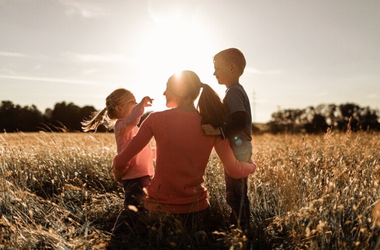 A person with a ponytail, seen from behind, kneels in a field of tall grass at sunset, sharing a special moment with two children. One child is standing and the other is sitting on the person's knee. They all appear to be smiling and enjoying the moment.