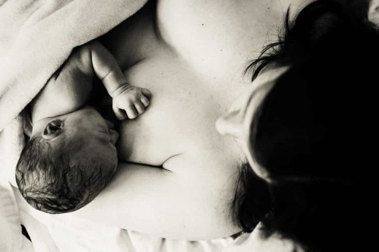 A newborn baby lies on an adult's chest, a tender moment that speaks to their intimate home birth story. The baby is partially covered by a blanket, and the dark-haired adult holds them close. The photo's black-and-white tones add timeless beauty to this precious scene.