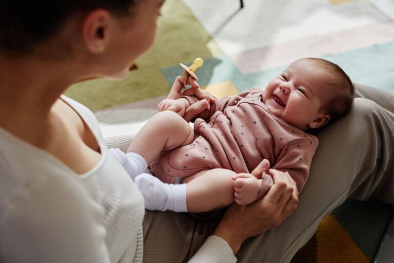 High angle view at a smiling baby wearing a pink polka dot onesie lies on an adults lap. The adult, in a white top, holds a pacifier and gazes affectionately at the baby. The scene unfolds in a room with a colorful carpet.