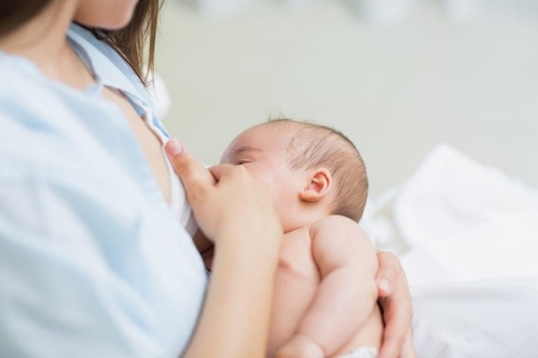 A woman is breastfeeding an infant, cradled in her left arm, nursing on her right breast. Despite common breastfeeding discomforts, the setting appears calm and intimate with soft lighting and a white background. The focus is on the bonding moment between mother and child.