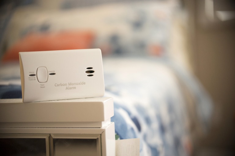 A carbon monoxide detector is placed on a piece of furniture in a bedroom. The background shows an out-of-focus bed with blue and white bedding.