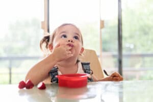 A 20-month-old toddler sits in a high chair at a table, eating from a red bowl. The child is wearing a white shirt and denim overalls. A few strawberries sit on the table, while large windows in the background frame a tranquil view of trees.