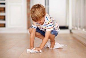 A young child is kneeling on a wooden floor, using a white cloth to clean up. The child is wearing a striped short-sleeve shirt and blue shorts. The background includes kitchen cabinets and appliances, showing how you can keep a clean house with kids involved in chores.