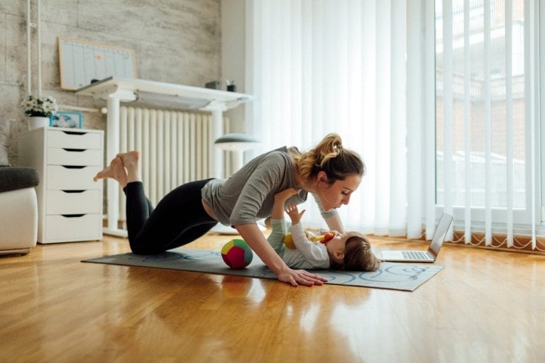 A woman performs a plank exercise on a yoga mat in a living room, playfully interacting with her baby lying on its back. The room features a white radiator, a desk, and large windows with vertical blinds letting in daylight. A colorful ball is on the mat. She embraces her loose skin while enjoying this moment.