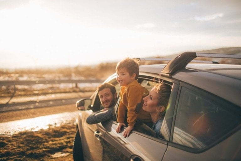 A man, a woman, and a young child are leaning out of the windows of a parked car on a sunny day. Traveling with kids seems to bring joy to all three as they smile together. The child is positioned between the man and woman, against a backdrop of a rural landscape with a blurred sunlit sky.