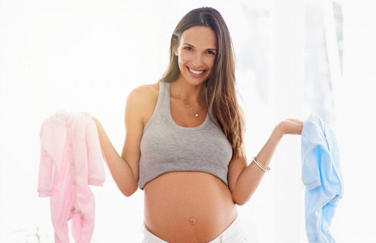 A pregnant woman smiles while holding up a pink baby outfit in her left hand and a blue baby outfit in her right hand, contemplating "How to Conceive a Boy." She is wearing a grey tank top and white pants. The background is softly lit and bright.