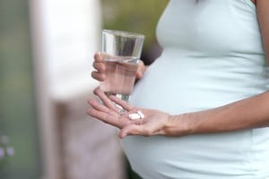 A pregnant person stands holding a glass of water in one hand and three prenatal vitamins in the other palm. The individual is wearing a light green sleeveless top. The background is blurry and suggests an outdoor setting.