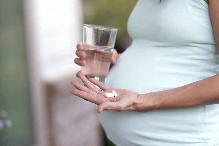 A pregnant person stands holding a glass of water in one hand and three prenatal vitamins in the other palm. The individual is wearing a light green sleeveless top. The background is blurry and suggests an outdoor setting.