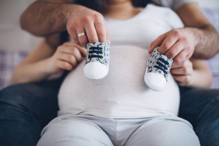 A pregnant woman sits, cradled by another person behind her. The person's hands hold a pair of baby shoes near the woman's belly. With her white shirt and light-colored pants, she radiates anticipation as she mentally prepares the birth center bag for the special day ahead.