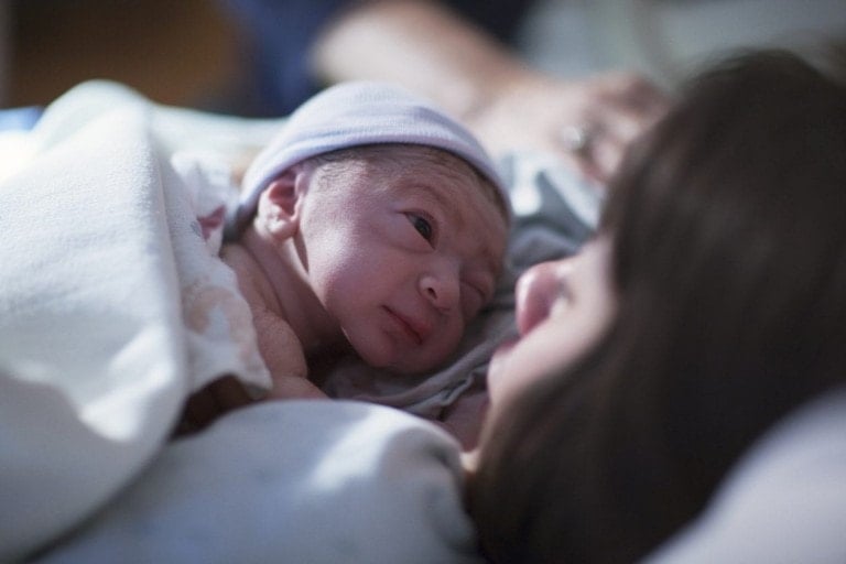 A newborn baby wrapped in a blanket and wearing a hat looks up at an adult who is lying down, likely the mother. The scene, part of her incredible journey to motherhood, appears to take place in a hospital or birthing center, with the baby resting gently on her chest.