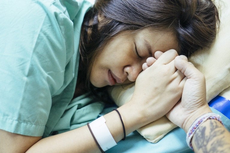 A person wearing a hospital gown lies in bed, holding hands with another. The individual in bed, adorned with a white hospital wristband, appears to be resting with eyes closed. Behind them are a pillow and a blue object, possibly reflecting on why a natural birth was chosen.